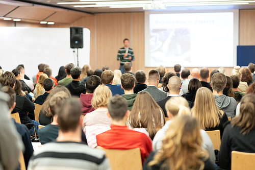 Speaker giving presentation in lecture hall at university. Participants listening to lecture and making notes.