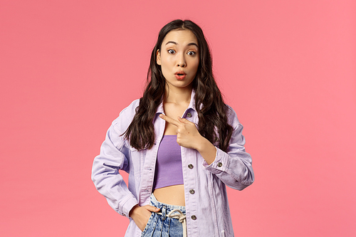 Portrait of surprised and curious young stylish female student pointing finger left and look amazed camera, asking question about interesting thing located there, pink background.