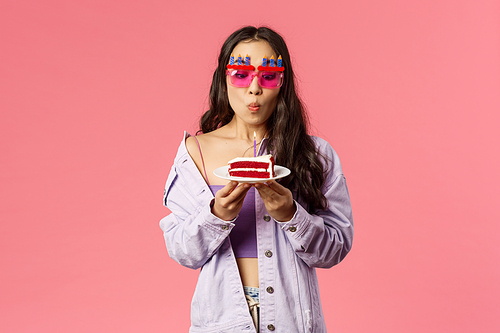 Celebration, holidays and people concept. Portrait of dreamy good-looking asian girl in b-day glasses, blowing lit candle on birthday cake, making wish, want dream come true, pink background.