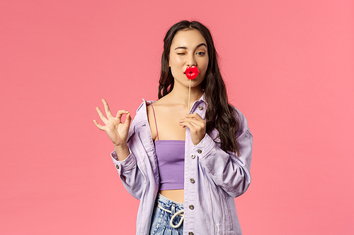 Holidays, lifestyle and people concept. Portrait of funny, sassy mixed-race girl in stylish outfit, show okay no problem sign, hold stick of big lips, wink cute camera, standing pink background.