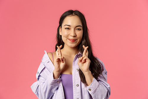 Close-up portrait of upbeat young asian girl hopefully awaiting for good results, cross fingers good luck, aspiring and anticipating great news, relish something, making wish, pink background.