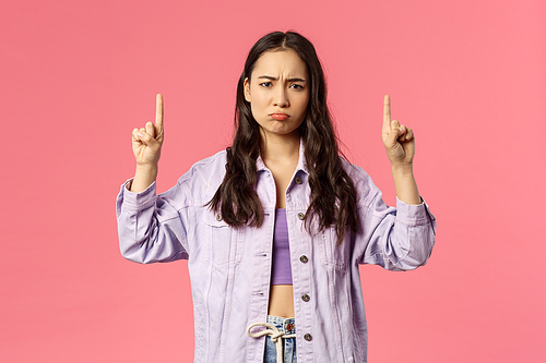 Portrait of uneasy, sulking unhappy asian girl in denim jacket over crop-top, complain unfair situation, pointing fingers up, look camera offended or gloomy, standing pink background.