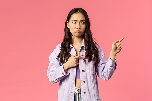 Attractive, gloomy mixed-race girl pointing fingers right, looking at something with regret and sigh, pouting displeased, jealous of missing good opportunity, want be there, pink background.