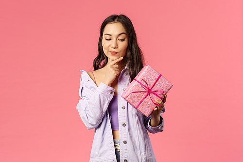 Portrait of curious enthusiastic young asian girl peeking at wrapped box, smirk and touching jaw as thinking about what inside of gift given by friend on birthday party, pink background.