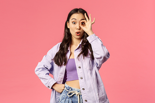 Portrait of surprised young woman seeing zero reasons not follow this page or buy product online shop, see something good, look through okay sign and fold lips say wow, pink background.