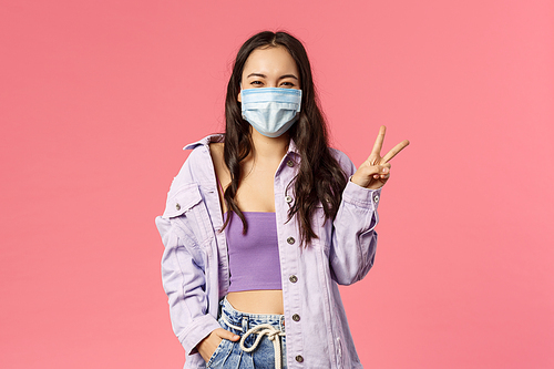 Covid19, quarantine, people concept. Optimistic, smiling happy girl in medical face mask, inside home, show peace sign, encourage stay safe during coronavirus pandemia outbreak, pink background.