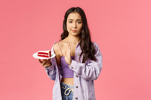 Portrait of skeptical young asian stylish slim girl refusing eat cake, smirk show top sign to dessert lying on plate, rejecting try it, disappointed with bad taste, stand pink background.