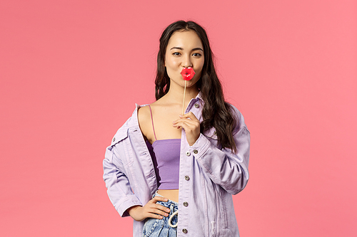 Holidays, lifestyle and people concept. Portrait of sassy attractive and stylish modern asian girl holding kissing lips stick and make confident expressing, showing her attitude, pink background.