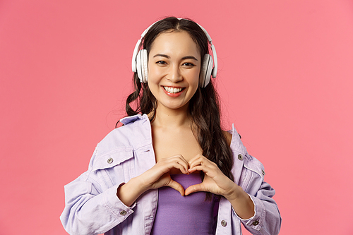 Close-up portrait of charming, lovely asian girl listening music in headphones, show heart gesture over chest, in love with her favourite artist, singer perform awesome new songs, pink background.