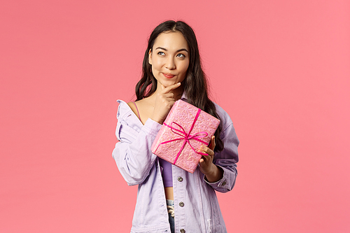 Portrait of happy thoughtful young asian girl receive wrapped box with gift, wonder what hidden inside, trying to guess, smile and look up thinking, standing pink background intrigued.