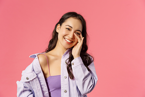 Lifestyle, fashion and beauty concept. Close-up portrait of carefree, smiling happy asian girl tilt head tender look camera, touching face, advertisement of skincare routine products, pink background.