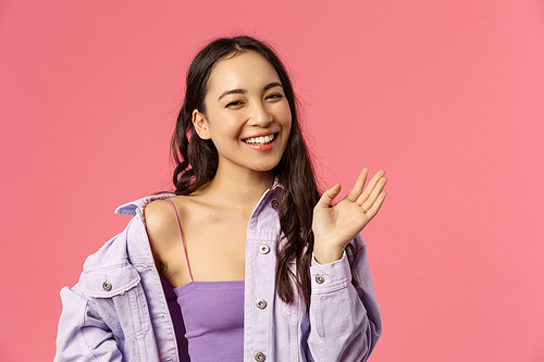 Close-up portrait of happy good-looking asian girl saying hi, waving hand casually, informal greeting sign, laughing and smiling friendly, welcome friend, greet hello, stand pink background.