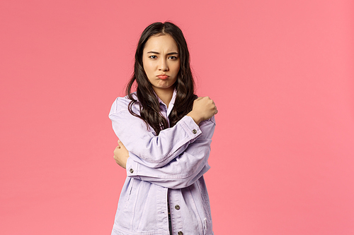 Portrait of silly and cute asian girl hugging herself and sulking, complaining on someone being rude and offensive to her, grimacing angry, waiting for apologies, standing pink background.