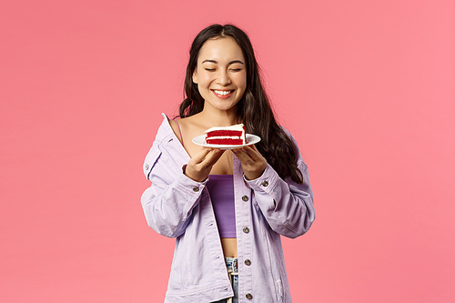 Portrait of happy, smiling and carefree asian girl close eyes and feel happiness holding favourite dessert, eating delicious cake, drop diet and enjoy sweets, standing pink background.