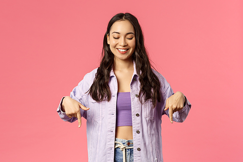 Portrait of stylish good-looking asian girl in denim jacket, crop-top, found great place to visit summer vacation, pointing and looking down with pleased beaming smile, pink background.