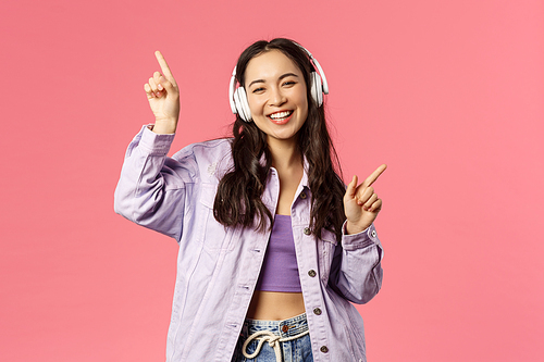 Lifestyle, technology and people concept. Portrait of carefree pretty young asian girl having fun, dancing joyfully, listening music in eadphones, raising hands up, singing along, pink background.