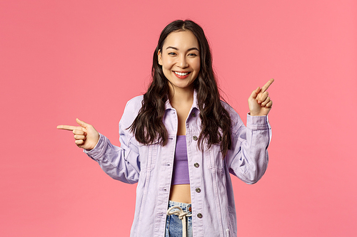 Portrait of stylish assertive cute korean girl giving choice, pointing fingers left and up, promote products, give advice where find useful links on online shops, internet stores, pink background.