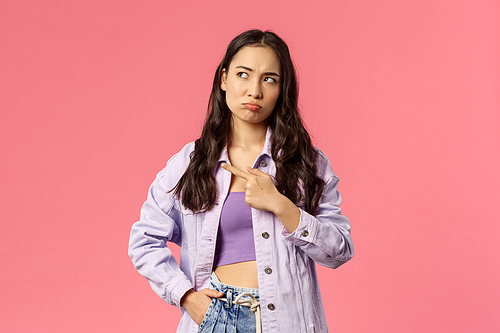 Portrait of complicated cute young korean girl in stylish outfit, frowning looking and pointing upper left corner, standing thoughtful, facing problem thinking of solution, pink background.