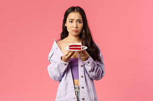 Portrait of gloomy sad cute asian girl just being dumped trying eat-out her sadness, holding piece cake, pouting and frowning upset, have uneasy feelings, grieving, pink background.