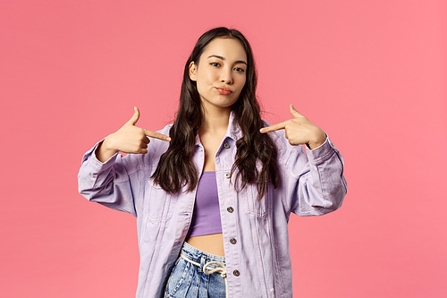 Portrait of cool and sassy stylish young woman pointing at herself with confident face, smirk and look camera as what you waiting for, pick me, bragging, being boastful, pink background.