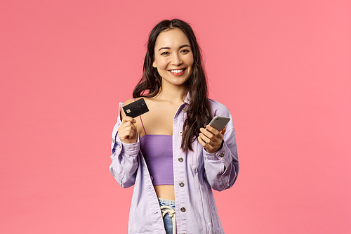 Portrait of young stylish girl, smiling pleased camera, using mobile phone and hold credit card to pay for online purchase, shop in internet, order food delivery, standing pink background.