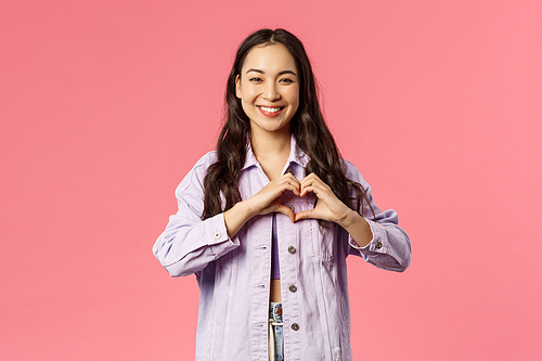 Portrait of cute, attractive asian girl in denim jacket, smiling as showing heart sign to confess love, being romantic, express sympathy or passionate feeling, falling for someone, pink background.