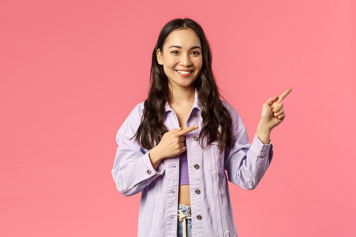 Portrait of cheerful asian girl, female student inviting people follow page, subscribe to page or visit link, pointing fingers right and smiling camera, standing happy over pink background.