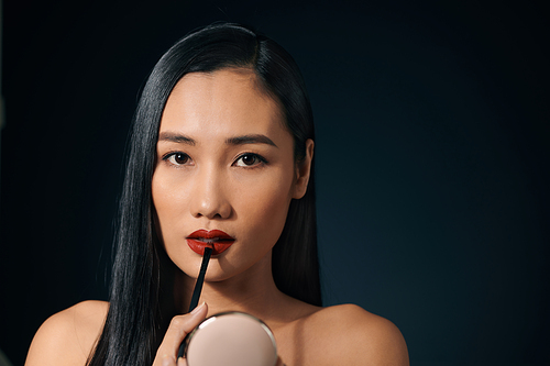 Young charming woman applying lipstick against black background.