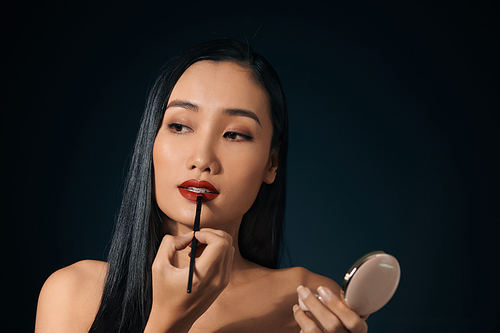 Young charming woman applying lipstick against black background.