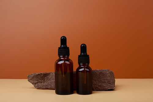 Two dark glass bottles with serum or oil for skin care next to stone on beige table against brown background. Cosmetic bottles with natural rough stone against dark background