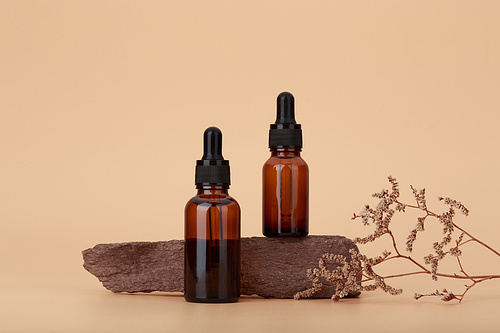 Two skin serum bottles next to natural stone and dry flower against beige background with copy space. Concept of luxury skin serum for anti aging treatment