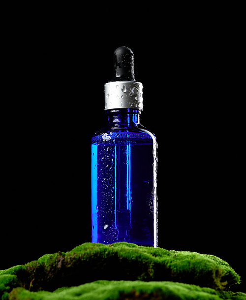 blue glass bottle with pipette stands on green moss, black background. Cosmetics SPA branding mockup