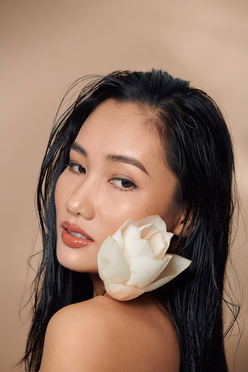 Woman got dark tan skin with long hair and holding flowers on beige background in white cropped top