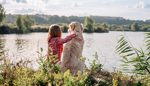 Child girl sitting and hugging golden retriever dog outdoors at the nature and looking at the lake. Teen kid with doggy pet resting close to river. Portrait from back