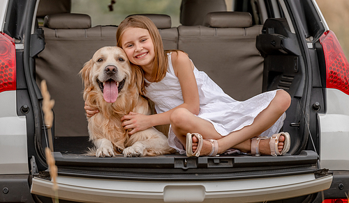 Little girl hugging golden retriever dog in car trunk and smiling. Cute child kid resting with doggy pet in vehicle during trip