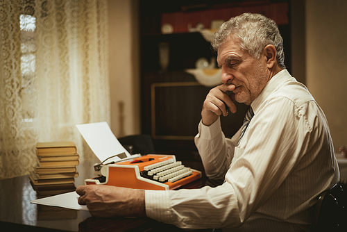 Pensive retro senior man sitting at the desk beside obsolete typewriter and thinking.