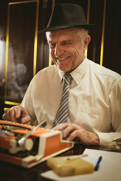 Retro senior man writer with hat on his had, smiling and typing on obsolete typewriter something amusing.