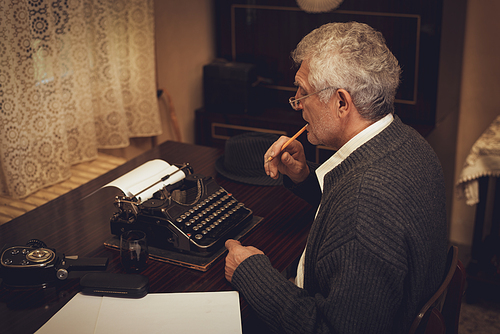 Retro senior man writer with glasses and pencil in his mouth sitting and looking on the text on obsolete typewriter.