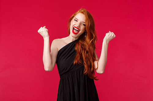 Portrait of successful young pleased redhead girl winning, feel pumped, raise fists up like champion, saying yes singing and make victory dance, triumphing in black dress over red background.