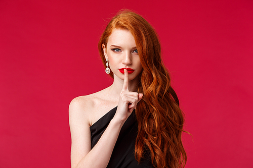 Shush lets keep it secret. Sassy and seductive good-looking redhead woman with red lipstick, earrings and black dress, hiding something, hush with smile on her face, tell be quiet, red background.
