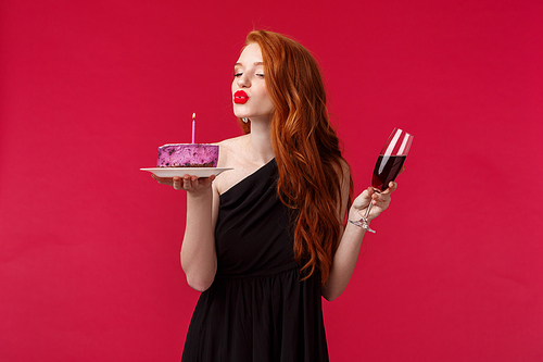 Portrait of elegant good-looking feminine redhead woman with red lipstick, evening makeup and black dress, celebrating birthday at party with red wine and b-day cake, blowing candle sensually.