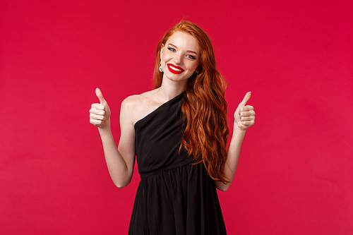 Fashion, luxury and beauty concept. Portrait of charming supportive young redhead woman in black elegant dress, makeup, smiling pleased show thumbs-up in approval or like, red background.