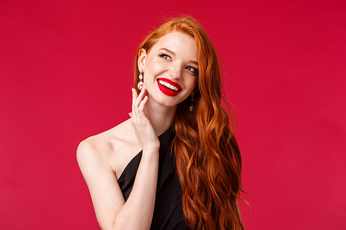 Close-up portrait of gorgeous luxurious and sophisticated young redhead rich female model attend fashion week or party in black evening dress, earrings and red lipstick, smiling look away dreamy.