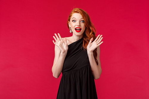 Fashion, luxury and beauty concept. Portrait of excited, overwhelmed young stunned redhead elegant woman in slim black dress, raising hands astonished and curious, look camera satisfied.