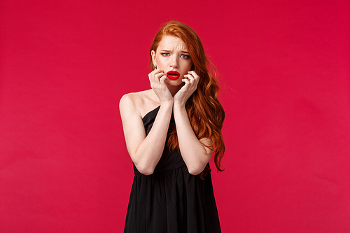 Fashion, luxury and beauty concept. Portrait of timid and scared ginger girl in black dress feel unsafe or insecure, touch face frowning frightened, look concerned and worried camera, red background.