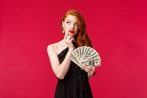 Luxury, beauty and money concept. Portrait of silly and thoughtful gorgeous redhead woman with beautiful ginger hair, wear black dress, pondering what buy on her prize, holding cash.