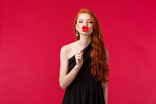 Portrait of feminine good-looking redhead woman in black stylish dress, holding fake lips cardboard over mouth as if give passionate kiss, looking at camera tender, stand red background.