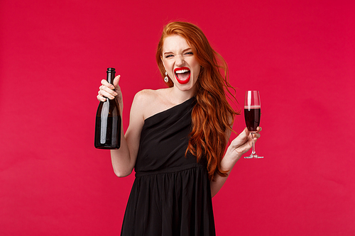 Portrait of excited sassy and carefree redhead drunk woman drinking from glass, holding champagne or wine bottle and shouting as having fun, party all night in elegant black dress, red background.