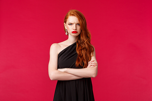 Portrait of angry and offended young gloomy frowning redhead girl in black evening dress, disappointed with someone, cross hands chest and grimacing irritatated, standing red background.