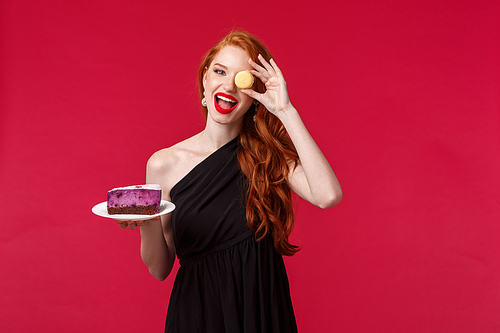 Portrait of gorgeous carefree redhead woman in slim black dress, hold macaron over eye funny smiling and cake on plate, party enjoy celebration of holiday, stand over red background.
